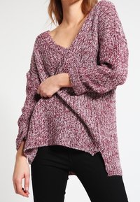 Gestrickter Pullover in Burgund- und Cremefarben, mit tiefem V-Ausschnitt, lockerem Schnitt und Seitenschlitzen. Das Material wirkt strukturiert und weich.