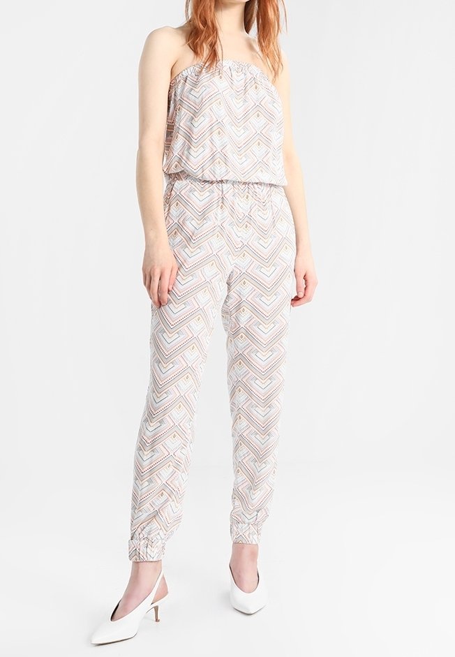 Femme portant une combinaison sans manches avec un motif chevron géométrique pastel et des chevilles élastiquées, assortie à des chaussures blanches à talons.