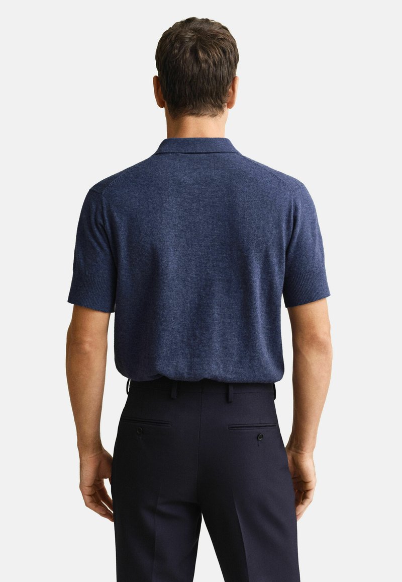 Homme portant un polo à manches courtes bleu marine, rentré dans un pantalon de ville noir, debout de dos sur un fond blanc.