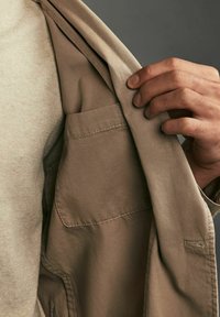 Veste khaki à texture lisse, dotée d'une poche poitrine et d'une coupe décontractée, portée par-dessus une chemise beige clair. Une main est visible tenant la veste.