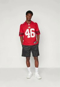 Camisa desportiva vermelha com números brancos "46" e riscas pretas; combinada com calções de rede pretos. O modelo usa meias brancas e sapatilhas.