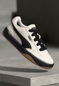Sapatilhas Puma com um cabedal sintético em creme, detalhes pretos, painéis texturizados, atacadores pretos e uma sola de borracha castanha.
