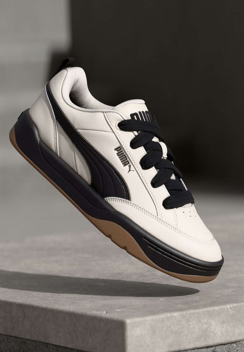 Sapatilhas Puma com um cabedal sintético em creme, detalhes pretos, painéis texturizados, atacadores pretos e uma sola de borracha castanha.