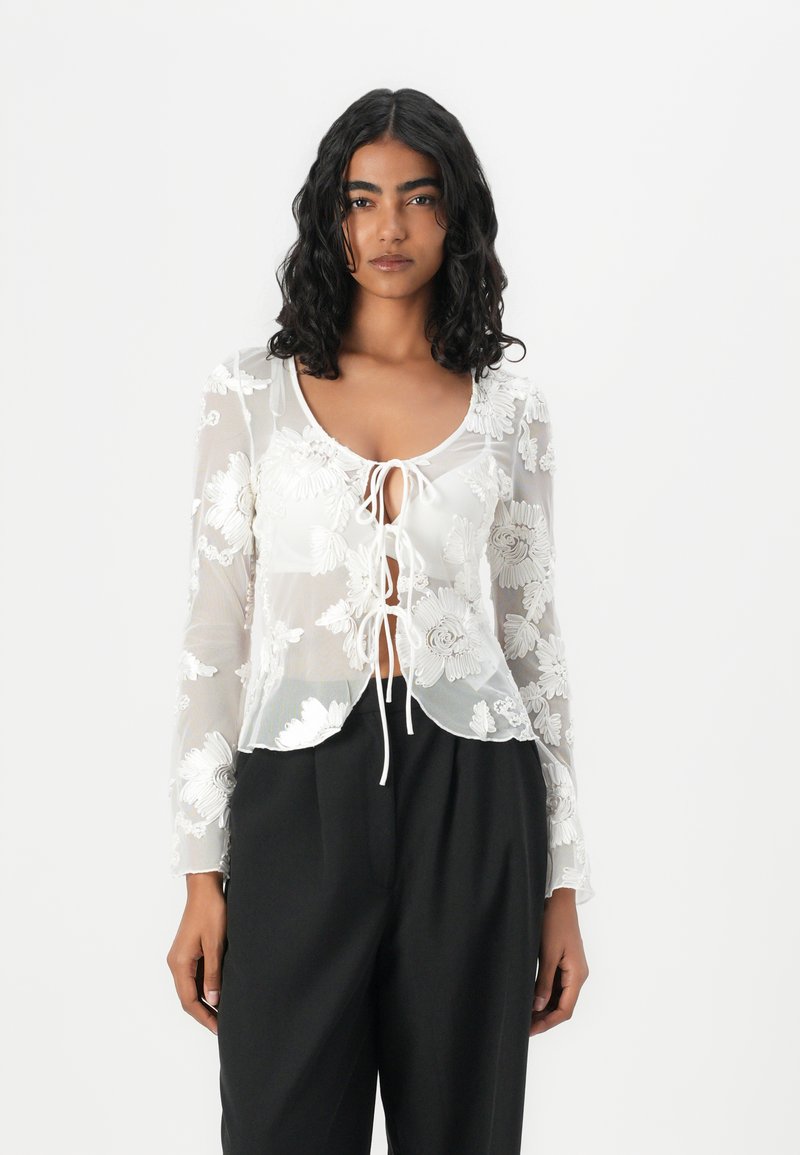Sheer white blouse with floral embroidery, scoop neckline, long sleeves, and drawstring front, paired with black trousers.