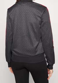 Svart jacka med texturerad tygstruktur som har ett subtilt upprepat logomönster, röda detaljer längs ärmarna och ribbade muddar och fåll.