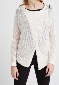 Pull en tricot blanc avec un design croisé lâche, présentant un motif texturé, un large col et deux poches avant.