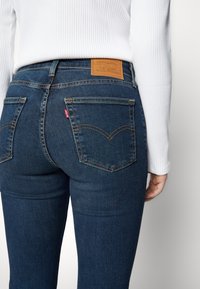 Gros plan d'une personne portant un jean denim bleu foncé avec des poches arrière et un patch de marque en cuir marron, associé à un haut blanc à manches longues côtelé.
