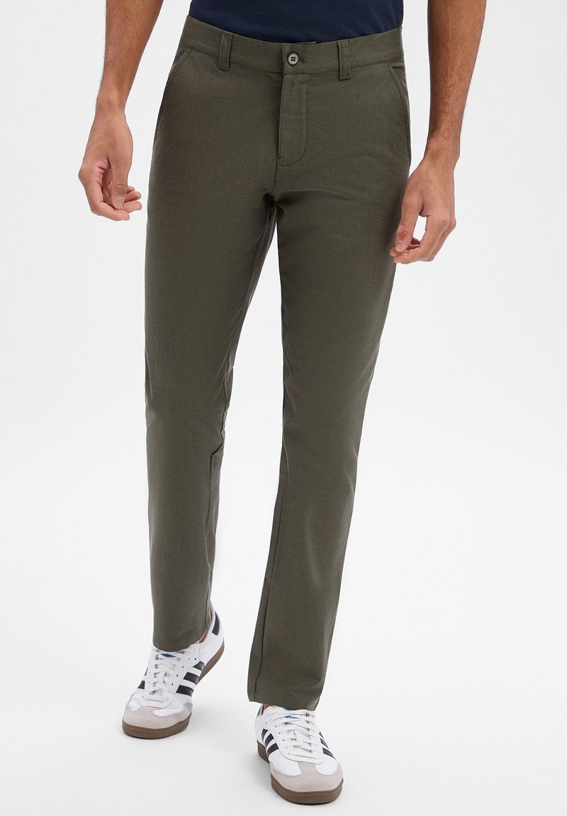 Homme portant un pantalon droit vert olive et des baskets blanches avec des rayures noires, debout devant un fond neutre.