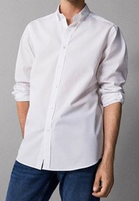 Man wearing a buttoned white long-sleeve shirt with sleeves rolled up and blue jeans, standing against a plain background.