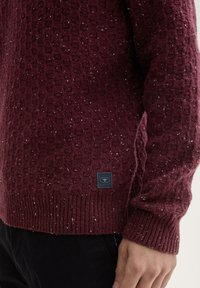 Bordeauxfarbener Strickpullover mit strukturiertem Muster und weißen Flecken. Mit gerippten Ärmelbündchen und Saum. Besitzt ein kleines schwarzes Etikett an der unteren Seite.