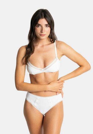 Femme aux longs cheveux foncés portant un soutien-gorge blanc et des sous-vêtements, debout avec un bras autour de la taille sur un fond clair uni.