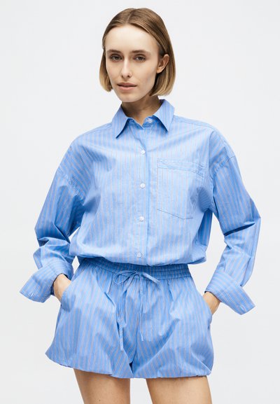 Γυναίκα που φοράει άνετο μπλε ριγωτό πουκάμισο με κουμπιά και ταιριαστό σορτς με κορδόνι, στέκεται με τα χέρια στις τσέπες μπροστά σε απλό φόντο.