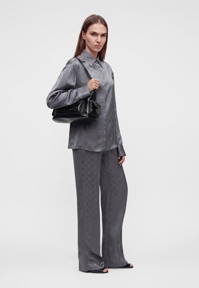 Femme portant une chemise et un pantalon larges en satin gris, tenant un sac à épaule noir, debout devant un fond clair uni.