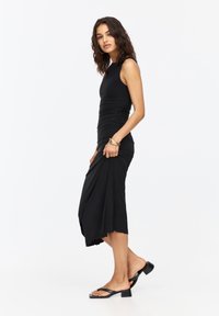 Schwarzes ärmelloses Kleid mit Raffung, enganliegendem Oberteil, fließendem Rock und knielanger Saumlinie. Kombiniert mit schwarzen, geschnürten Blockabsatzsandalen.