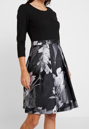 Frau trägt ein schwarzes Kleid mit dreiviertel Ärmeln und einem knielangen Rock mit schwarz-weißem Blumenmuster.
