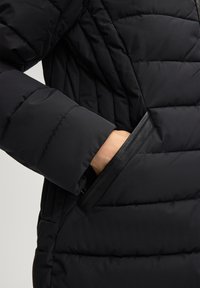 Gewatteerde zwarte jas met een doorgestikt patroon, voorzien van een leren accent rondom de zak en een nauwsluitend manchetdesign.