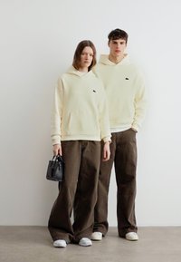 Δύο άτομα ντυμένα με κρεμ φούτερ με κουκούλα και καφέ παντελόνια φαρδιάς γραμμής στέκονται μπροστά σε ένα λευκό τοίχο· το ένα κρατά μια μικρή μαύρη τσάντα χειρός.