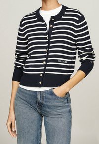 Cardigan a righe blu navy e bianche con colletto arrotondato, chiusura con bottoni e polsini a costine. Presenta il logo "HILFIGER" in bianco.