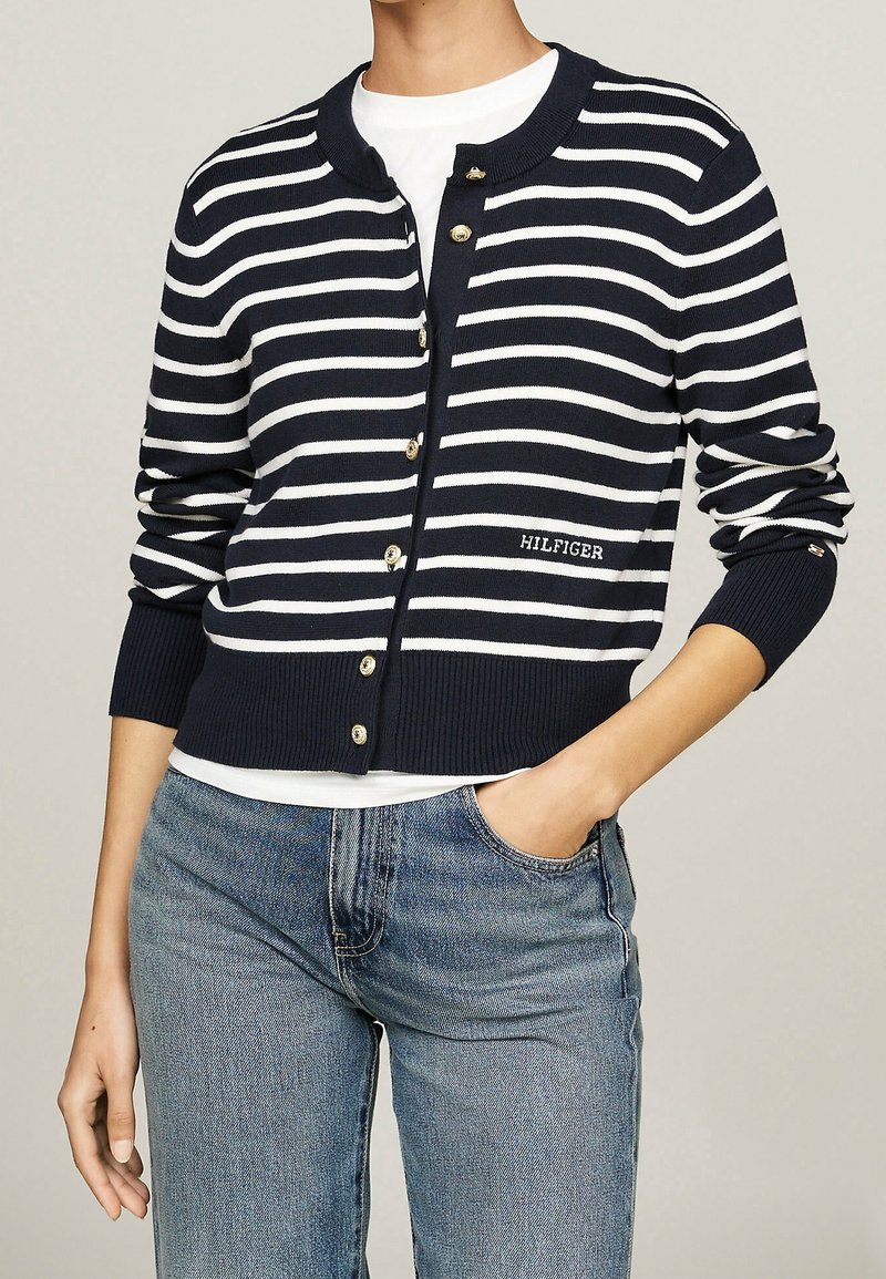 Cardigan a righe blu navy e bianche con colletto arrotondato, chiusura con bottoni e polsini a costine. Presenta il logo "HILFIGER" in bianco.