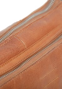 Läderväska med en slät, tan-finish och synliga sömmar. Har en silvrig metallzipper längs toppen. Inredningen är fodrad med tyg.