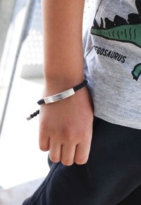 Zwarte gevlochten armband met een zilveren metalen plaatje gegraveerd met "LEef JE VERHAAL," gedragen om de pols van een kind. Casual grijs shirt is gedeeltelijk zichtbaar.
