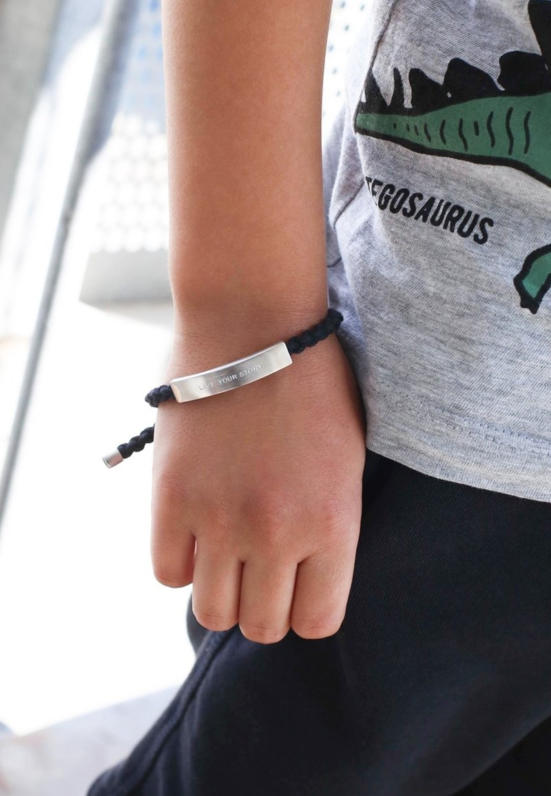 Zwarte gevlochten armband met een zilveren metalen plaatje gegraveerd met "LEef JE VERHAAL," gedragen om de pols van een kind. Casual grijs shirt is gedeeltelijk zichtbaar.