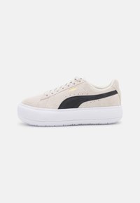 La sneaker en suède beige présente une rayure noire, une semelle en caoutchouc blanc et des détails perforés. La marque dorée sur le côté ajoute un contraste.