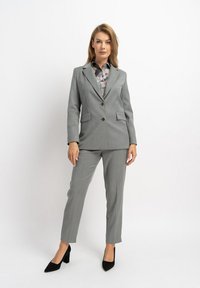 Grau maßgeschneiderter Anzug mit einem Blazer mit zwei Knöpfen und aufgesetzten Taschen, kombiniert mit passenden Hosen. Das Outfit wird durch ein mehrfarbiges Hemd ergänzt.