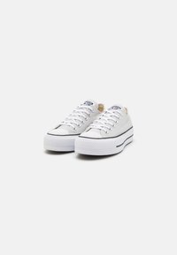 Weiße Canvas-Sneaker mit niedrigem Design, schwarzer Gummisohle und schwarzem Streifen; ausgestattet mit weißen Schnürsenkeln und einem kleinen Logo auf der Zunge.