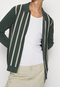 Grön stickad cardigan med vertikala ränder i beige och vitt, med en dragkedja fram, krage och ribbade ärmslut. Bärs över en vit tank top.