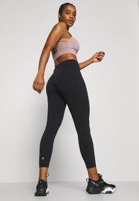 Schwarze Leggings mit hohem Bund und geschwungenen Nähten. Kombiniert mit einem hellrosa, taillierten Sport-BH mit offenem Rücken. Schwarze Sportschuhe.