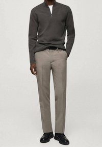 Polo gris à fermeture éclair avec une texture lisse, associé à un pantalon beige clair sur mesure. Chaussures noires complètent la tenue. Fond neutre.