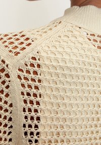 Sweter z dzianiny w kolorze kremowym, z otwartym splotem. Prezentuje teksturowany, siatkowy wzór z prążkowanym kołnierzem i ramionami.
