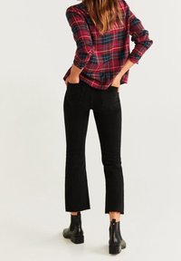 Vrouw in een rood en marineblauwe geruite shirt, zwarte cropped jeans met rafels aan de onderkant en zwarte enkellaarsjes, staand met haar handen in de achterzakken.