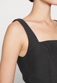 Robe noire texturée avec des bretelles larges et un décolleté carré. Couture visible le long des côtés et du corsage.