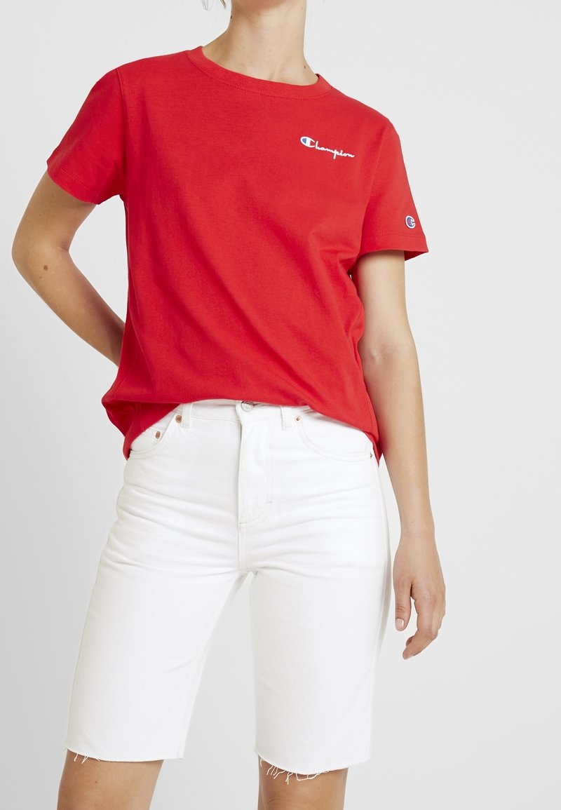 Rød bomulds t-shirt med korte ærmer og rund halsudskæring, der har et lille broderet logo. Sammen med hvide denim shorts.
