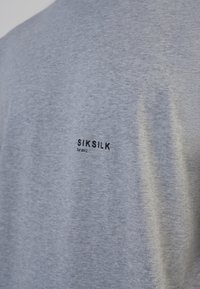 Pelēks T-krekls izgatavots no kokvilnas materiāla. Aizmugurē ir melns drukāts logo ar uzrakstu "SIKSILK" un "Est 2012" horizontāli. Vienkāršs dizains, īsas piedurknes.