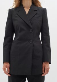 Femme portant un blazer noir ajusté avec deux boutons, des revers crantés et des manches longues, montrée du cou jusqu'aux hanches.