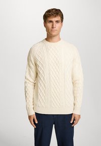 Crèmekleurige kabelgebreide trui met een ronde hals, voorzien van gestructureerde patronen en geribbelde mouwen en zoom. Gedragen over marineblauwe broek.