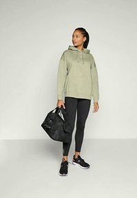 Haut à capuche vert olive avec une poche sur le devant, associé à un legging noir et des chaussures de sport noires. Portant un sac de sport noir avec des anses.