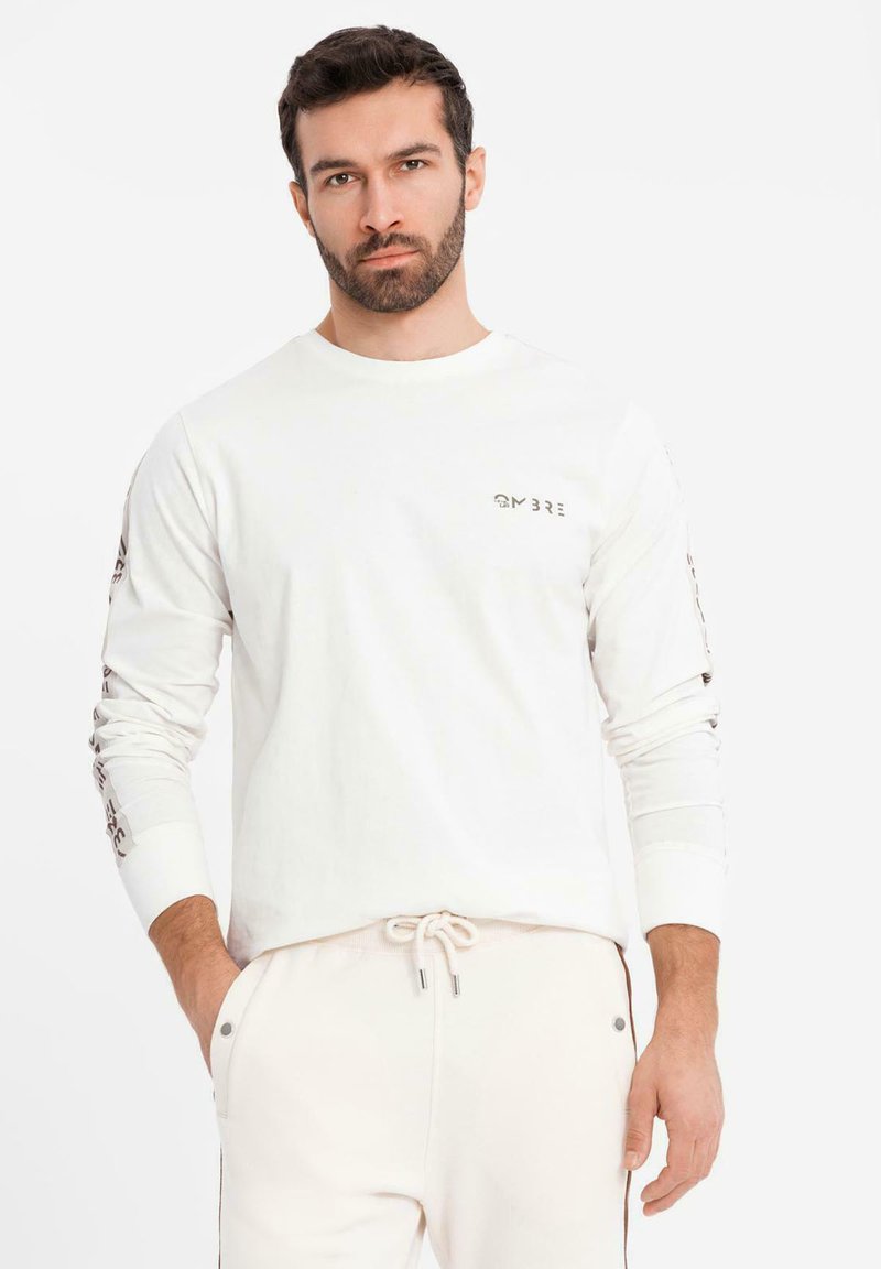 Muž s plnovousem, oblečený v bílé košili s dlouhými rukávy, na hrudi a rukávech s textem "OMBRE", v kombinaci se světle zbarvenými kalhotami na šňůrku, s rukama v kapsách.