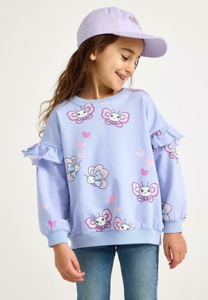 Ung pige smiler, iført en lys lilla kasket og en pastelblå sweatshirt med smilende sommerfugl- og hjerteprint, kombineret med blå jeans.