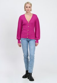 Fuchsia ribbet cardigan med dyb V-hals og lange, puffede ærmer. Har træknapper og bæres med lyseblå jeans og sorte ankelstøvler.