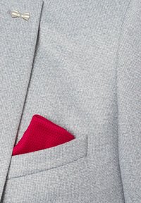Grauer gemusterter Blazer mit einer silbernen Fliege und einem rosa Taschentuch mit perforierter Textur, ordentlich in der Brusttasche platziert.
