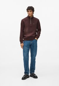 Barna, cipzáras nyakú pulcsi, finom logóval a mellkason, kék farmerral és fekete cipővel párosítva, egy sima fehér háttér előtt állva.