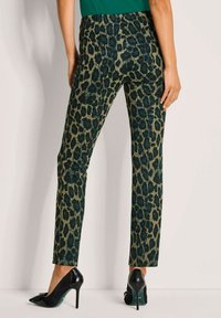 Vrouw in groene en zwarte luipaardprint broek, gecombineerd met zwarte hoge hakken, staand tegen een lichte achtergrond.