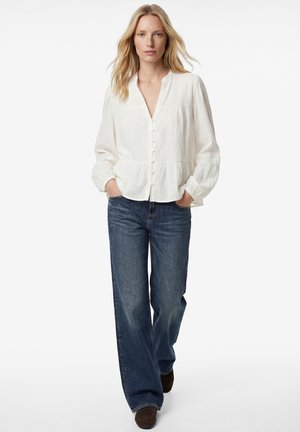 Frau mit blondem Haar, die eine weiße langärmelige, geknöpfte Bluse und weit geschnittene blaue Jeans trägt, die Hände in den Taschen, geht nach vorne.