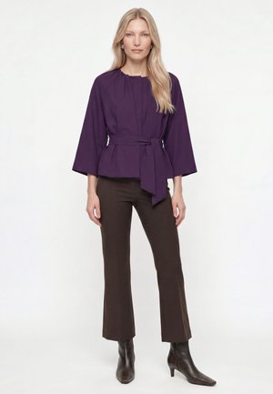 Woman with long blonde hair wearing a purple belted jacket, dark brown pants, and dark brown ankle boots standing against a white background.
