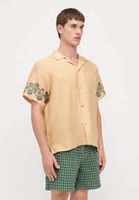 Camisa de lino de manga corta, de color amarillo claro, con bordados en espiral verdes en las mangas y botones sutiles marrones; combinada con pantalones cortos a cuadros verdes.