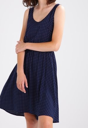 Mujer con vestido azul marino sin mangas con lunares blancos, de pie con un brazo cruzado sobre el cuerpo frente a un fondo liso.